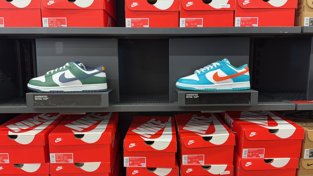 Two Nike Dunk Low colorways displayed on an outlet shelf, with stacked orange Nike shoeboxes below